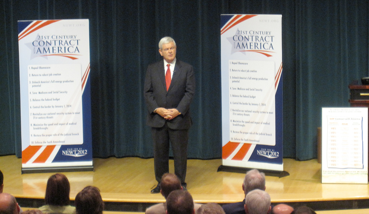 American politician Newt Gingrich, in a dark blue suit and red tie, stands on stage between two large vertical banners describing his 'contract with American'