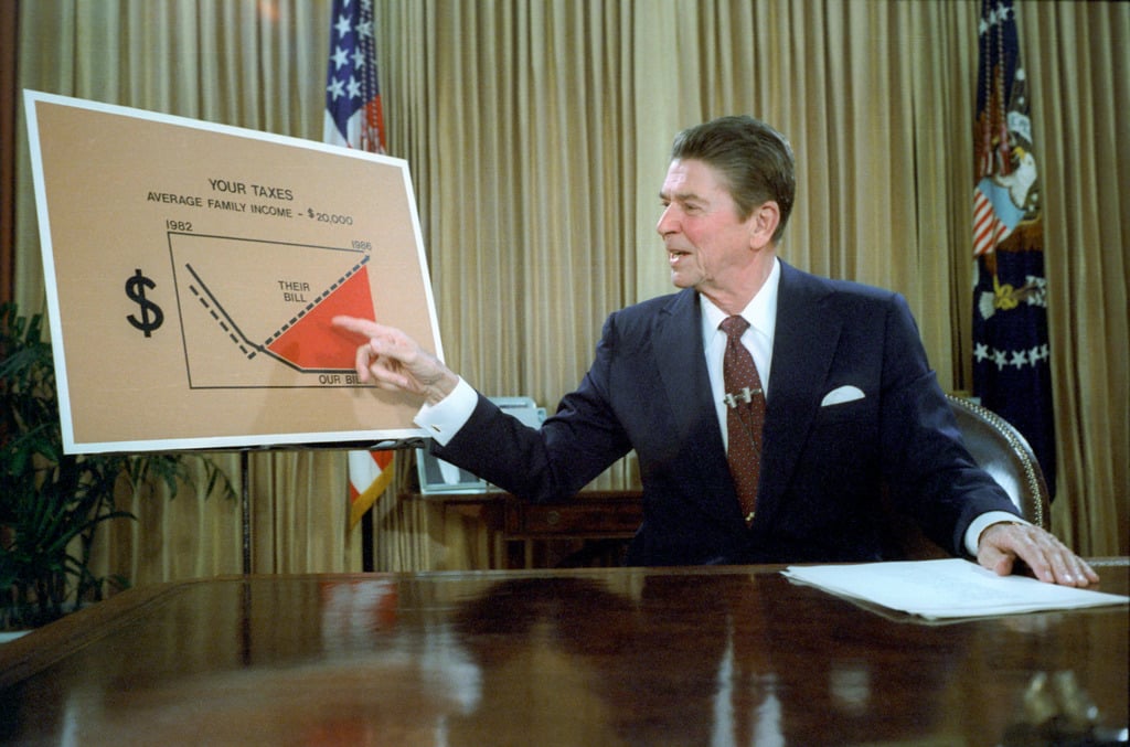 President Ronald Reagan behind a desk points at a large chart illustrating government expenditure. He is addressing the nation from the oval office on tax reduction. From the US Government NARA & DVIDS Public Domain Archive.
