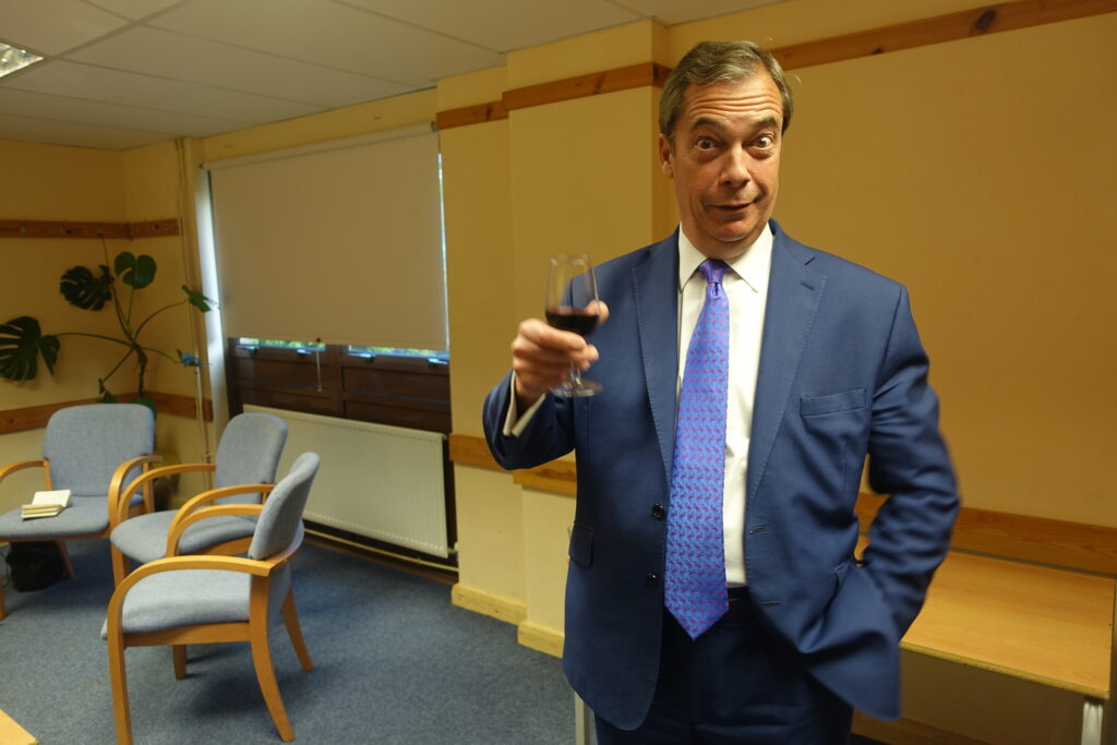Nigel Farage raises a glass of wine in a BBC green room in May 2017. He's gurning characteristically.