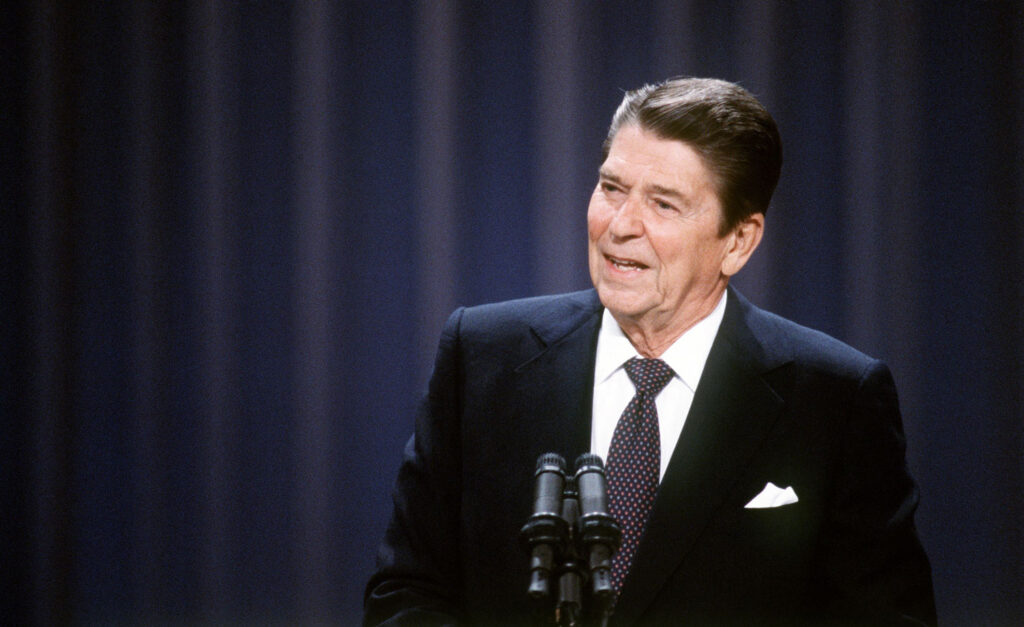 President of the USA Ronald Reagan against a blue curtained background making a speech behind two press microphones