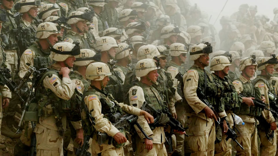 A large crowd of American soldiers in full uniform with helmets and weapons against a dusty background