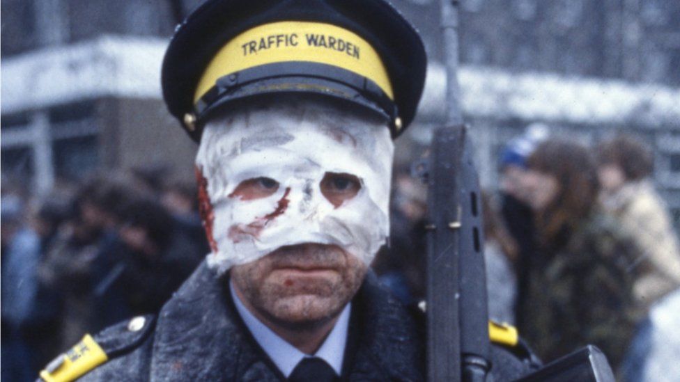 Famous still from 1984 film 'Threads' about the aftermath of a nuclear war in Britain. A man in a traffic warden's uniform has a bloody bandage with crude eye-holes covering almost his whole face. A rifle at his shoulder, dazed expression on his face.