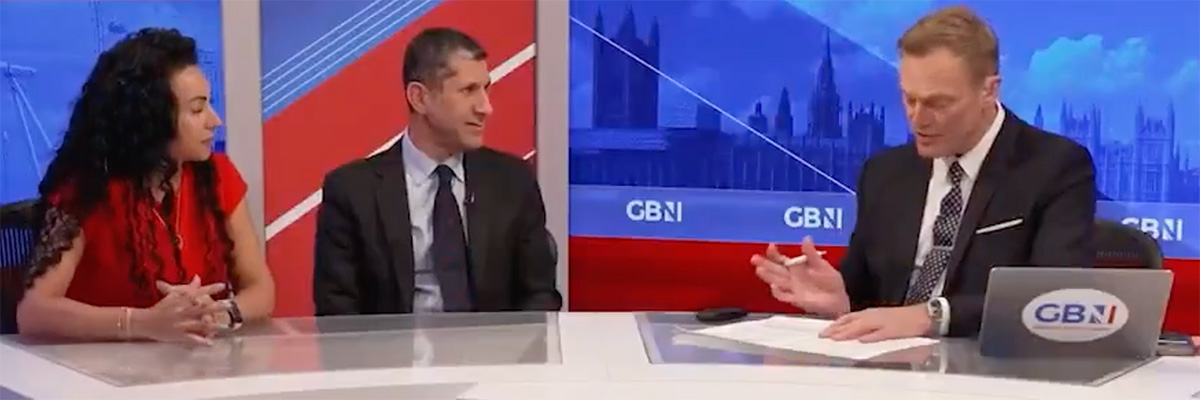 In a TV News studio two guests - a man and a woman - sit behind the presenter's desk alongside the news presenter. Colours and bright red and blue